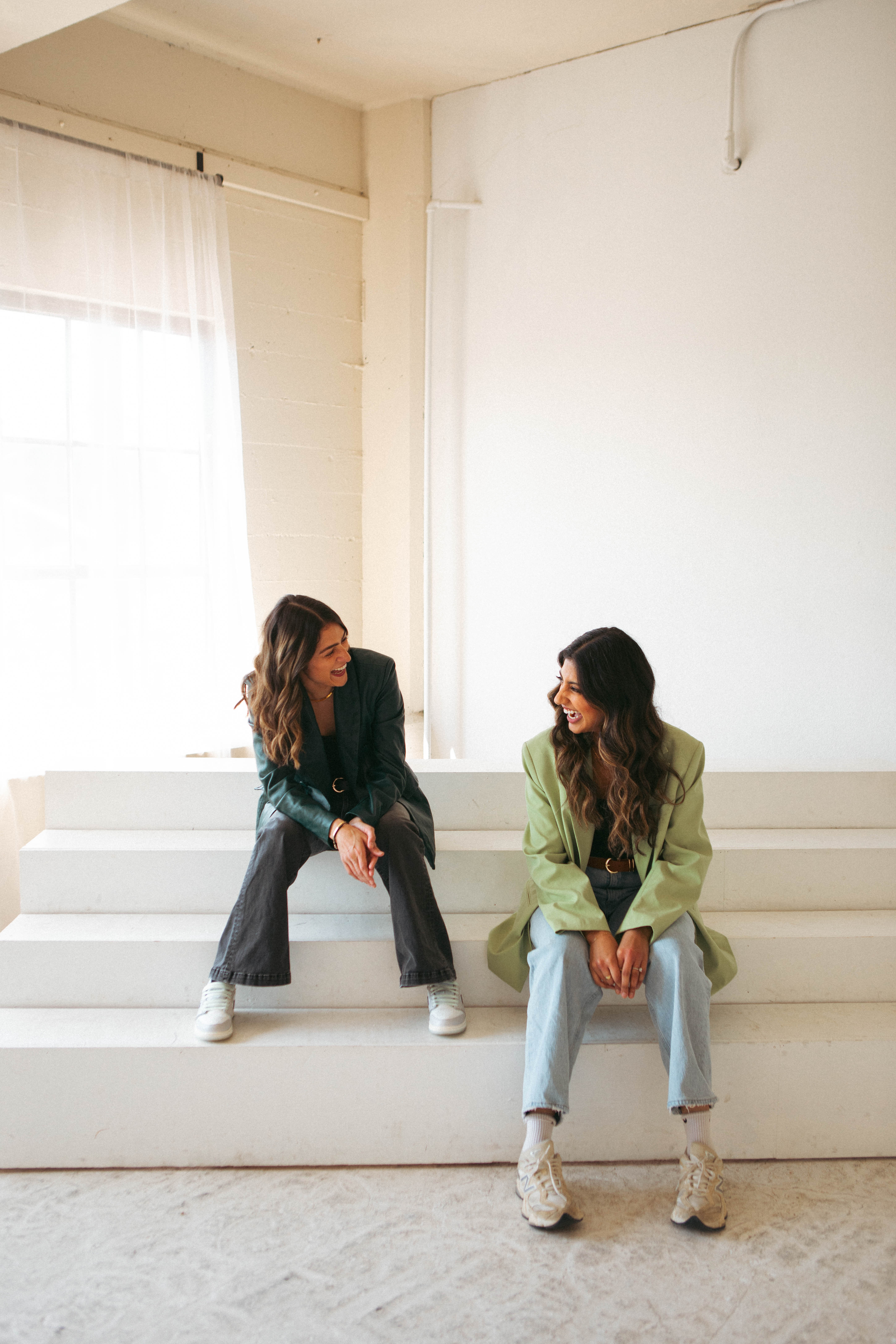 Purvi and Karen in green blazers laughing together on white studio steps