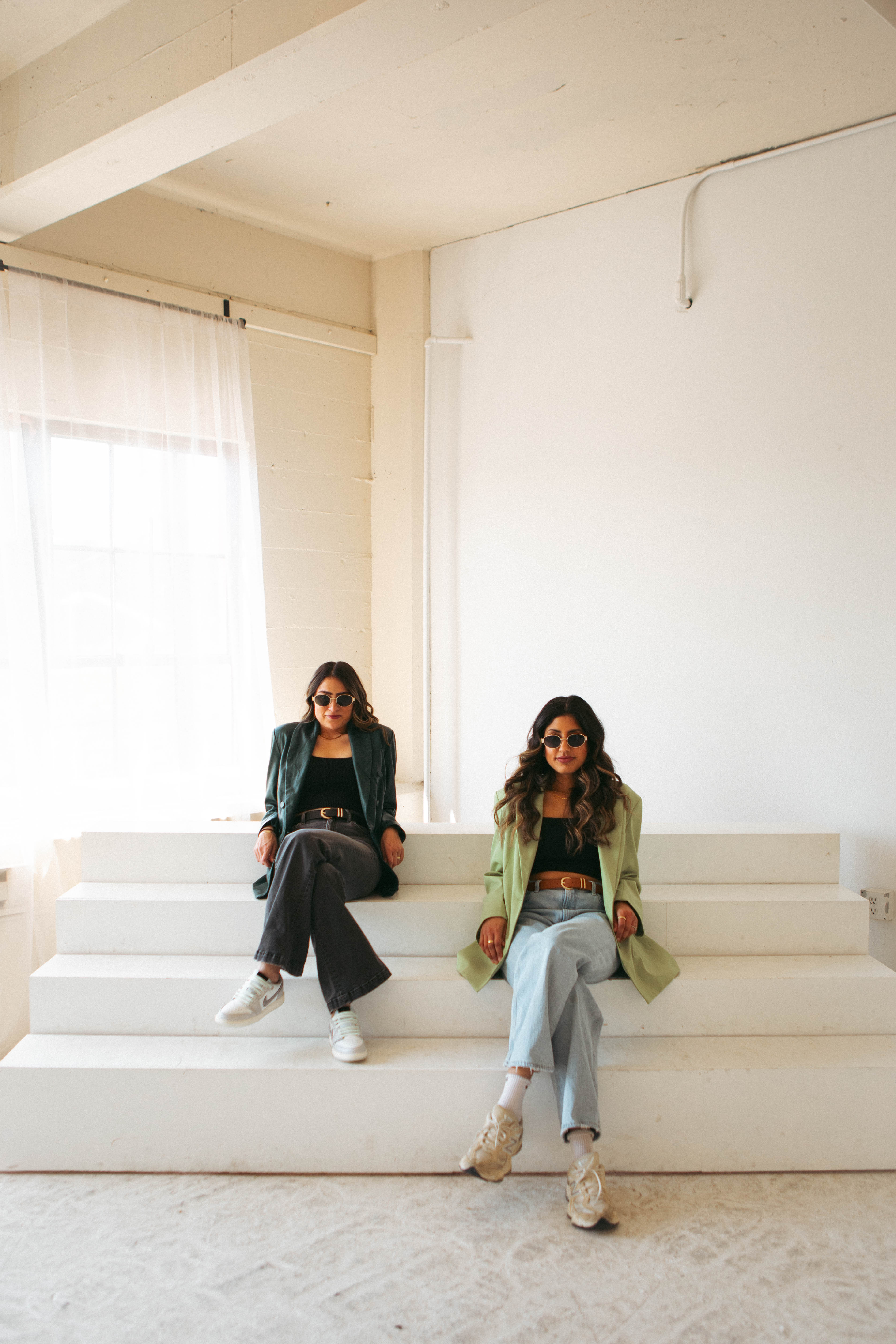 Purvi and Karen in sunglasses and green blazers, sitting on white studio steps