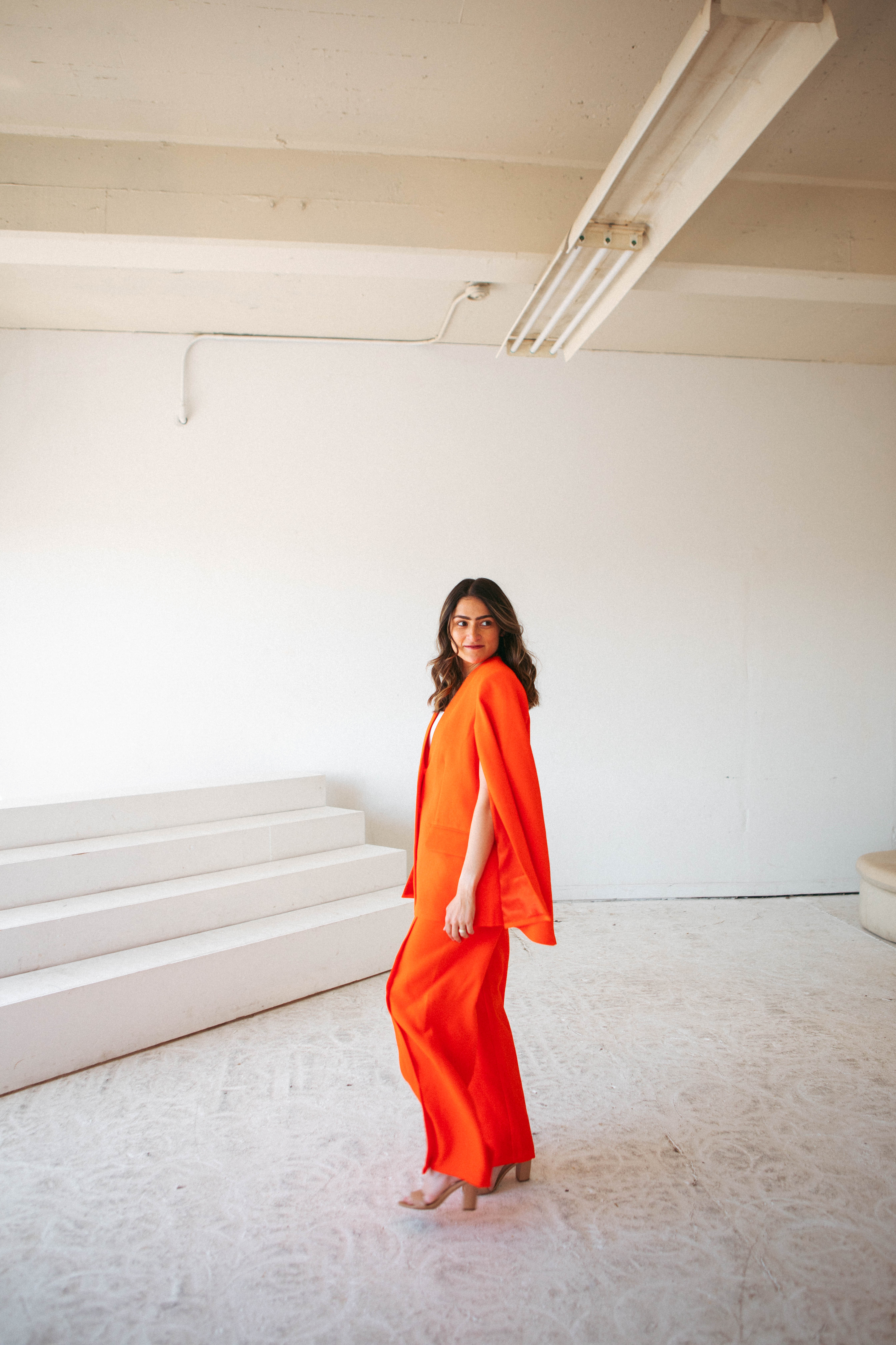 Karen Desai in an orange cape suit, glancing over her shoulder in a white studio