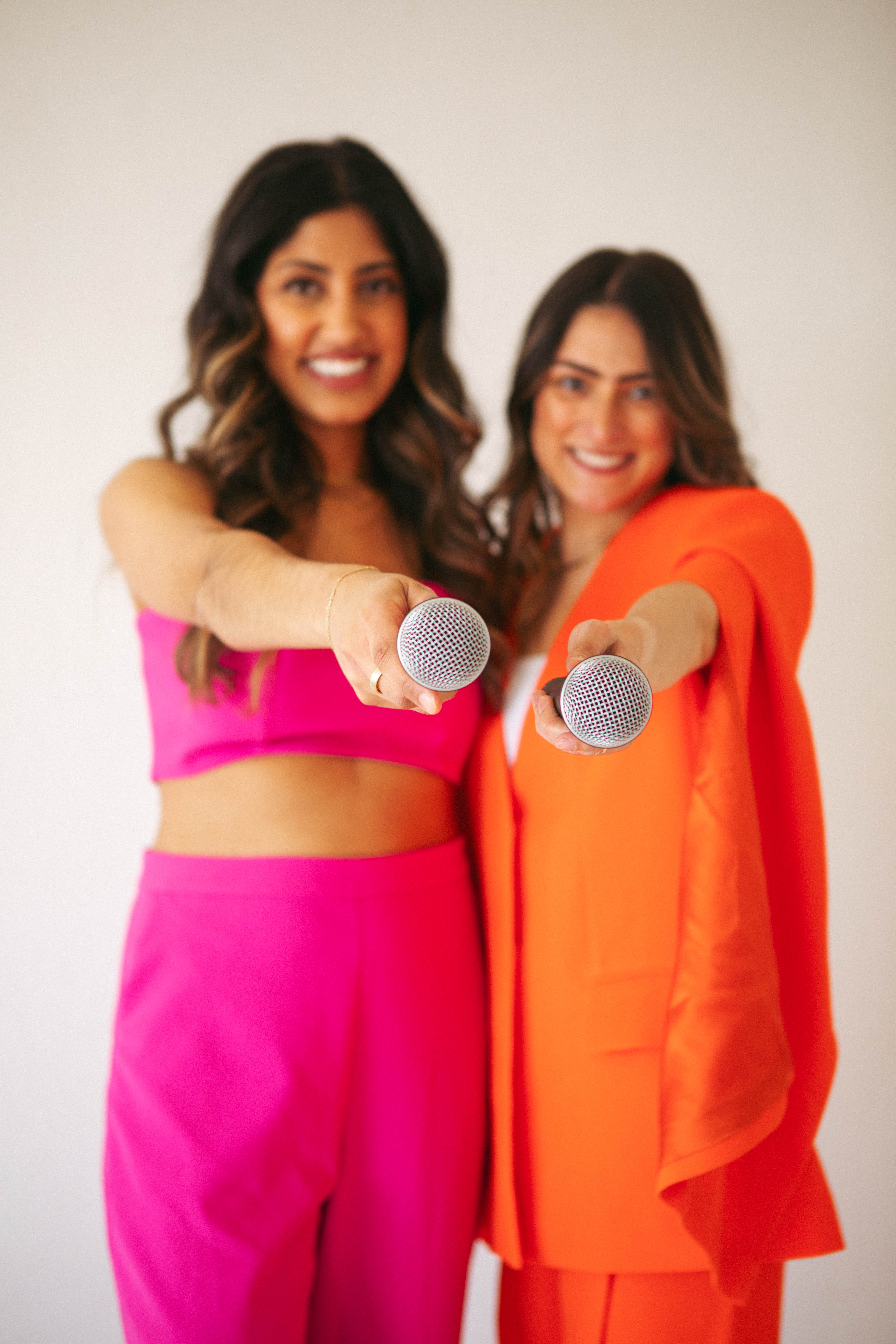 Purvi and Karen in bright pink and orange, holding microphones out toward the camera
