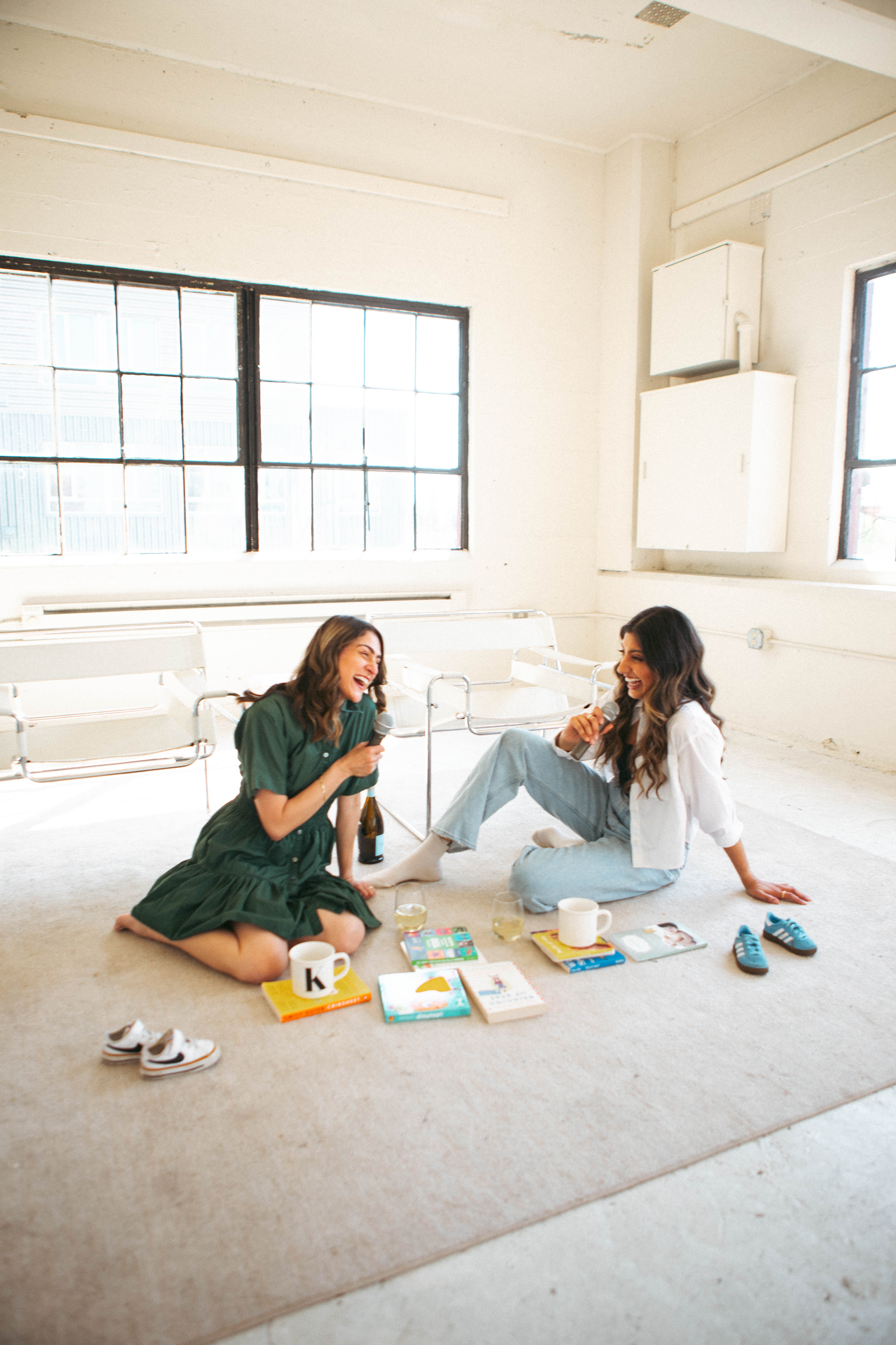 Purvi and Karen laughing with microphones on the studio floor, surrounded by parenting books, mugs, and tiny kid shoes